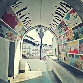 A beautifully painted archway features colorful comic-style artwork, with scenes depicted in vibrant panels on the walls. Through the archway, a view of a cityscape with quaint buildings and a tall palm tree is visible. The setting appears charming and artistic.