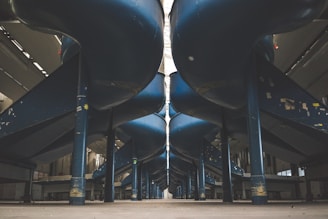 An industrial setting with large, blue cylindrical slides or ducts running parallel above the ground, supported by strong vertical beams. The area is spacious, with high ceilings and muted lighting, giving a sense of vastness.