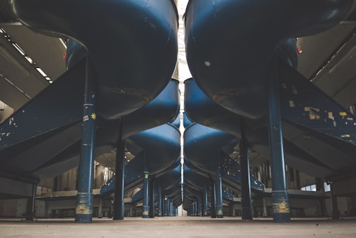 An industrial setting with large, blue cylindrical slides or ducts running parallel above the ground, supported by strong vertical beams. The area is spacious, with high ceilings and muted lighting, giving a sense of vastness.