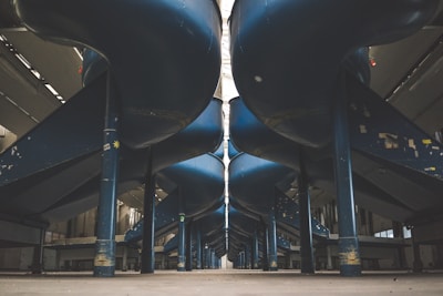 An industrial setting with large, blue cylindrical slides or ducts running parallel above the ground, supported by strong vertical beams. The area is spacious, with high ceilings and muted lighting, giving a sense of vastness.