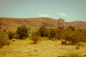 A vast desert landscape with dry, sandy ground and scattered vegetation. A metal water tank stands elevated on a rusted structure to the right, set against the backdrop of low, barren mountains. Sparse trees and bushes are visible, and the sky is clear with a few clouds.