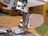 Close-up of a sewing machine needle and presser foot area showing detailed metallic parts and a plastic section on the right. The background is slightly out of focus, hinting at a domestic setting or workspace environment.