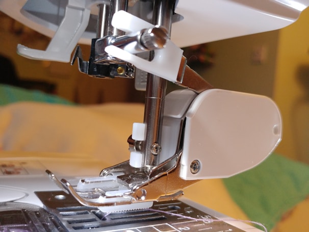 Close-up of a sewing machine needle and presser foot area showing detailed metallic parts and a plastic section on the right. The background is slightly out of focus, hinting at a domestic setting or workspace environment.