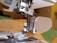 Close-up of a sewing machine needle and presser foot area showing detailed metallic parts and a plastic section on the right. The background is slightly out of focus, hinting at a domestic setting or workspace environment.
