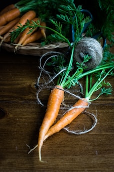 A close-up of two fresh, orange carrots with green tops on a wooden surface. They are tied together with rustic twine, suggesting a farm or rustic setting. In the background, there is a basket filled with more carrots, and a spool of twine is visible.
