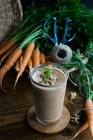 A glass filled with a creamy brown smoothie, topped with walnut pieces and a sprig of green garnish, is placed on a decorative wooden coaster. Around it, there are fresh carrots with greens, a pair of blue-handled scissors, and a spool of twine on a wooden table.