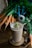 A glass filled with a creamy brown smoothie, topped with walnut pieces and a sprig of green garnish, is placed on a decorative wooden coaster. Around it, there are fresh carrots with greens, a pair of blue-handled scissors, and a spool of twine on a wooden table.