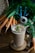 A glass filled with a creamy brown smoothie, topped with walnut pieces and a sprig of green garnish, is placed on a decorative wooden coaster. Around it, there are fresh carrots with greens, a pair of blue-handled scissors, and a spool of twine on a wooden table.
