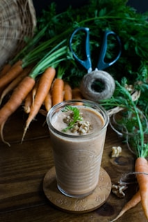 A glass filled with a creamy brown smoothie, topped with walnut pieces and a sprig of green garnish, is placed on a decorative wooden coaster. Around it, there are fresh carrots with greens, a pair of blue-handled scissors, and a spool of twine on a wooden table.