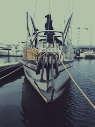 A boat docked at a marina, featuring a large anchor at the front. The boat is moored with ropes to the dock, surrounded by calm waters. Other boats and cranes are visible in the background, suggesting an industrial or commercial harbor setting.
