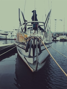 A boat docked at a marina, featuring a large anchor at the front. The boat is moored with ropes to the dock, surrounded by calm waters. Other boats and cranes are visible in the background, suggesting an industrial or commercial harbor setting.