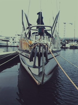 A boat docked at a marina, featuring a large anchor at the front. The boat is moored with ropes to the dock, surrounded by calm waters. Other boats and cranes are visible in the background, suggesting an industrial or commercial harbor setting.