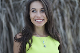 A woman with long brown hair and a bright smile is wearing a lime green top. She has a necklace with a blue pendant and a round medallion. The background appears to have out-of-focus plants or grass.