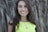 A woman with long brown hair and a bright smile is wearing a lime green top. She has a necklace with a blue pendant and a round medallion. The background appears to have out-of-focus plants or grass.