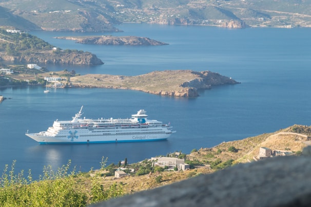 A large cruise ship is navigating through calm waters of a coastal area, surrounded by rugged hills and sparsely vegetated landscapes. Small clusters of buildings are visible near the shoreline, and the mountains in the background create a sense of depth and scale.