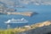 A large cruise ship is navigating through calm waters of a coastal area, surrounded by rugged hills and sparsely vegetated landscapes. Small clusters of buildings are visible near the shoreline, and the mountains in the background create a sense of depth and scale.