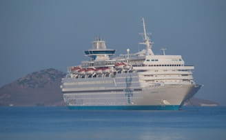 A large cruise ship is anchored in calm blue waters. The ship is white with multiple decks and visible lifeboats. In the background, there is a small, rocky island. The clear sky and gentle waves suggest tranquility.