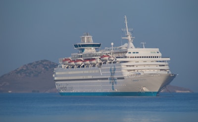A large cruise ship is anchored in calm blue waters. The ship is white with multiple decks and visible lifeboats. In the background, there is a small, rocky island. The clear sky and gentle waves suggest tranquility.