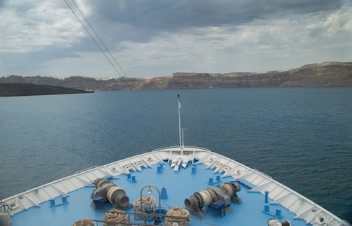 A ship's bow is pointed toward the rocky coastline of an island under a cloudy sky. The sea is calm and extends to the horizon, framed by the rugged landscape in the distance. Various equipment and ropes are visible on the deck, with a flagpole at the front.