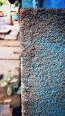 A textured concrete wall is close-up in the foreground, displaying a rugged surface with small stones and rough patches. In the background, an urban street scene is visible with blurred sidewalks and buildings, suggesting depth and perspective.