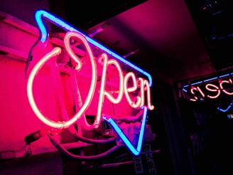 A glowing neon open sign casting a warm pink light against a dark wall at night.