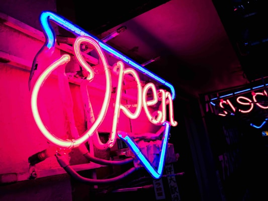 A glowing neon open sign casting a warm pink light against a dark wall at night.