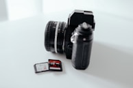 A black vintage-style camera is positioned on a smooth, white surface. Next to it are two memory cards, one of which is partially inserted into a slot. The camera features a prominent lens with silver and black detailing, and the memory cards have red and black labels.