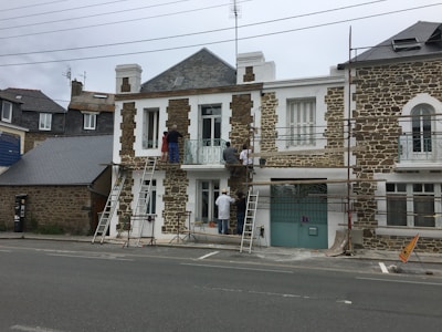 A group of people is working on the facade of a stone and white house, using scaffolding and ladders. The building has a classic architectural style with large windows and a wrought iron balcony. There are construction materials and tools visible on the ground.