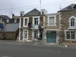 A group of people is working on the facade of a stone and white house, using scaffolding and ladders. The building has a classic architectural style with large windows and a wrought iron balcony. There are construction materials and tools visible on the ground.