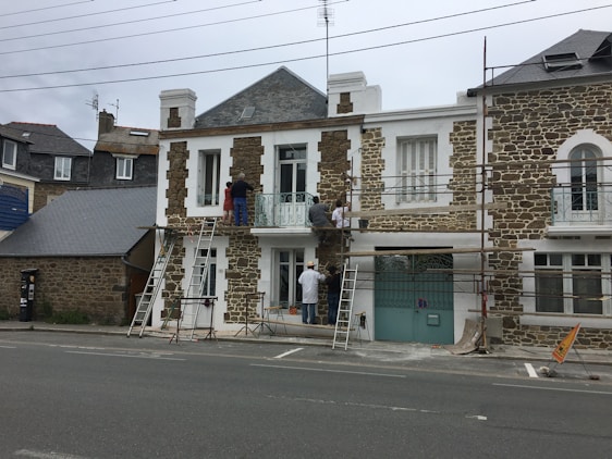 A group of people is working on the facade of a stone and white house, using scaffolding and ladders. The building has a classic architectural style with large windows and a wrought iron balcony. There are construction materials and tools visible on the ground.