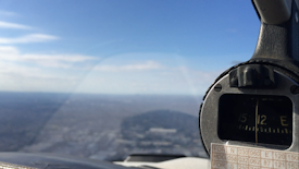 The photograph captures the view from inside a cockpit with a focus on an aircraft instrument panel or gauge. The background shows an expansive, clear sky with a hint of clouds over a distant landscape.