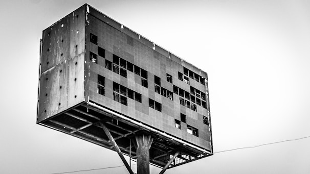 A large metal structure resembling a billboard or industrial installation is elevated on a sturdy support. The surface is dotted with rectangular openings and is weathered and rusty, giving it an aged appearance. The sky in the background is overcast, adding to the industrial and somewhat desolate feel.