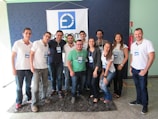 A group of eleven people is standing together indoors, posing for a photo. All individuals are wearing name tags and appear to be smiling, suggesting a positive atmosphere. They are positioned in front of a blue and white banner with a logo, which suggests a professional or event setting. The group is a mix of men and women, and most are wearing casual attire consisting of jeans and t-shirts or polos.