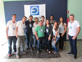 A group of eleven people is standing together indoors, posing for a photo. All individuals are wearing name tags and appear to be smiling, suggesting a positive atmosphere. They are positioned in front of a blue and white banner with a logo, which suggests a professional or event setting. The group is a mix of men and women, and most are wearing casual attire consisting of jeans and t-shirts or polos.