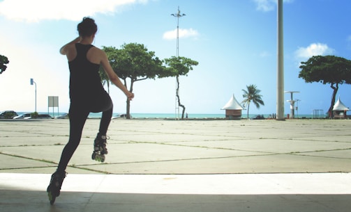 A person wearing dark athletic clothes and rollerblades is performing a skating maneuver on a wide, open plaza. There are green trees, a single palm tree, and white structures in the background, with the ocean visible under a blue sky.