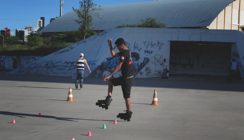 A person roller-skates in an outdoor skate park, performing a jump over a series of small colorful cones. The background features graffiti-covered walls and a few other skaters. The space is open, with some greenery and an industrial structure visible.