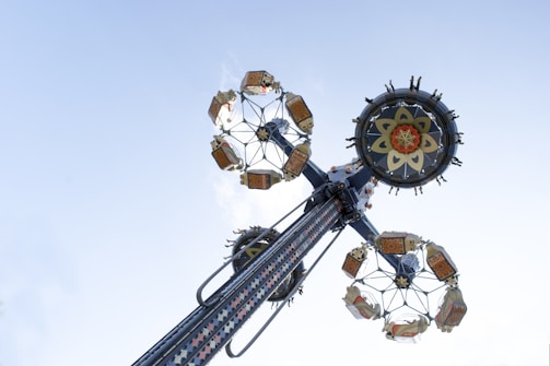 A tall amusement park ride extends into the sky, with multiple seats attached to large rotating wheels decorated with intricate patterns. The ride appears to spin around a central axis, with people seated at the outer edges.