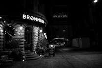 A dimly lit street corner with a vintage-style brick building displaying the illuminated sign 'BR&Oslash;NNUM'. The facade is adorned with classic lantern-style lights and potted plants. There is an archway leading to a darker, empty street visible in the background.