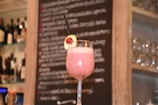 A pink smoothie garnished with a slice of banana and a raspberry is served in a tall glass with a black straw. The background features a blurry chalkboard menu with wine bottles and glassware shelves on either side.