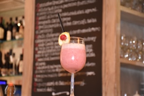 A pink smoothie garnished with a slice of banana and a raspberry is served in a tall glass with a black straw. The background features a blurry chalkboard menu with wine bottles and glassware shelves on either side.