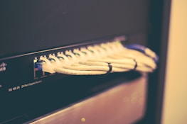 A network switch with multiple Ethernet cables plugged into its ports, positioned horizontally and bundled together with cable ties. The switch casing has visible brand markings and numeric labels near the ports.