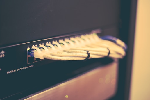 A network switch with multiple Ethernet cables plugged into its ports, positioned horizontally and bundled together with cable ties. The switch casing has visible brand markings and numeric labels near the ports.