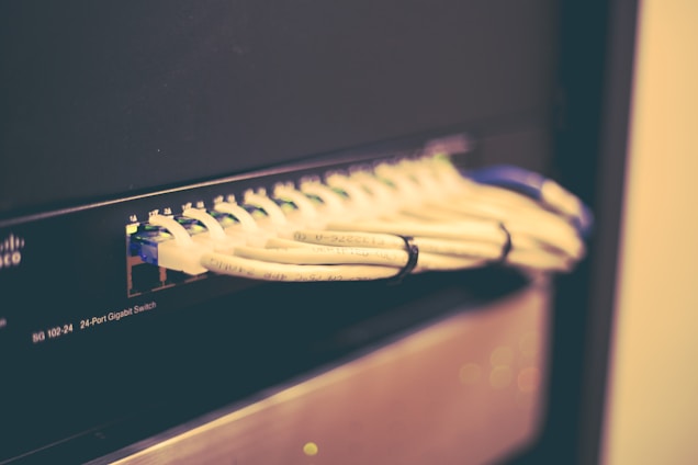 A network switch with multiple Ethernet cables plugged into its ports, positioned horizontally and bundled together with cable ties. The switch casing has visible brand markings and numeric labels near the ports.