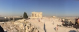 Ancient ruins stand prominently in an open, arid landscape with numerous tourists walking around. The sky is clear and blue, and the sun casts long shadows on the ground. The background reveals a sprawling cityscape.