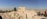 Ancient ruins stand prominently in an open, arid landscape with numerous tourists walking around. The sky is clear and blue, and the sun casts long shadows on the ground. The background reveals a sprawling cityscape.