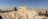 Ancient ruins stand prominently in an open, arid landscape with numerous tourists walking around. The sky is clear and blue, and the sun casts long shadows on the ground. The background reveals a sprawling cityscape.