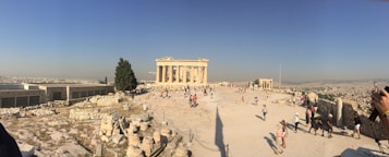 Ancient ruins stand prominently in an open, arid landscape with numerous tourists walking around. The sky is clear and blue, and the sun casts long shadows on the ground. The background reveals a sprawling cityscape.