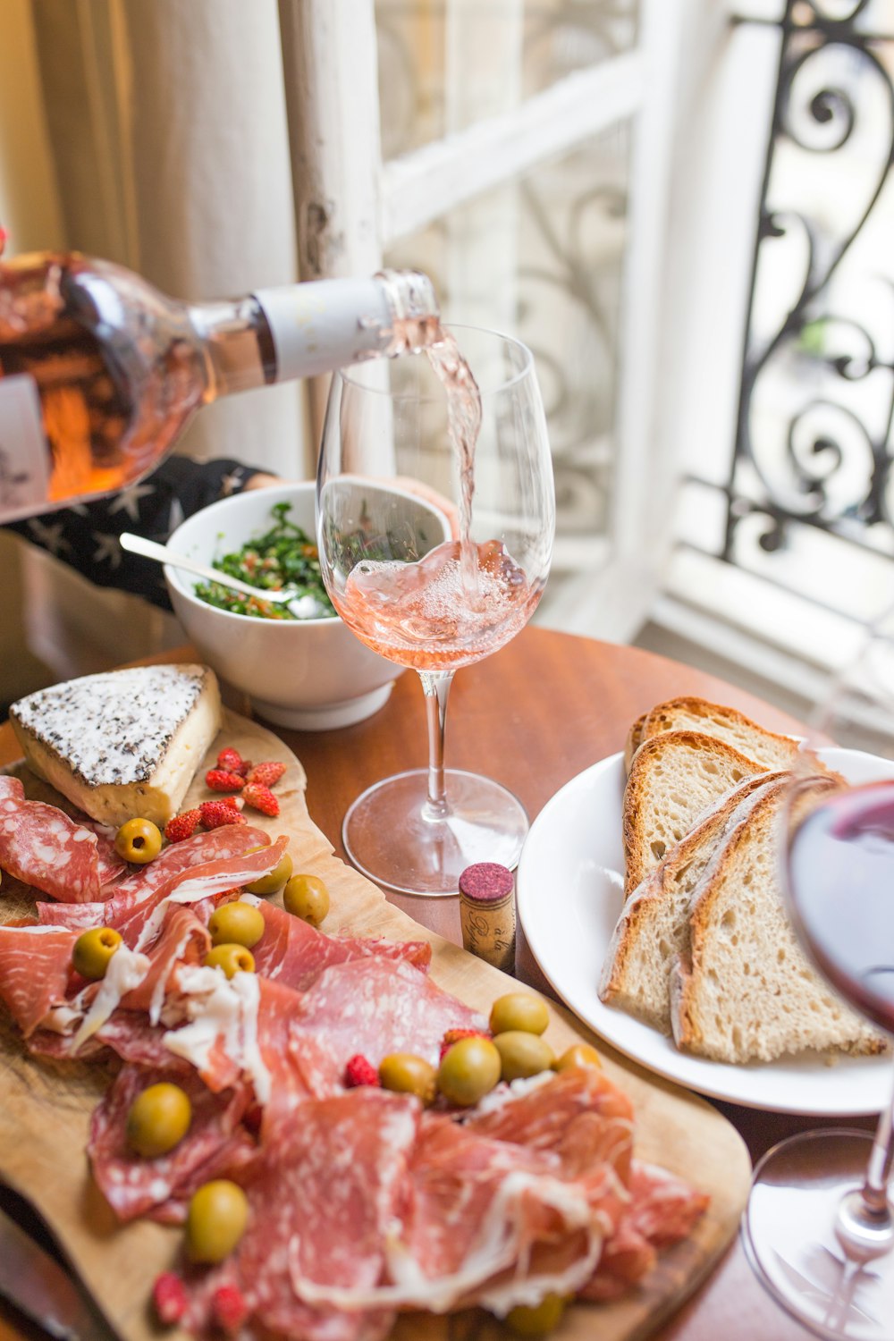 A person pouring rose into a wine glass, a cheese board with brie and salami, and bread on a table in Paris