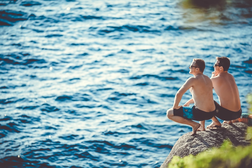 two men sitting on cliff in front of body of water