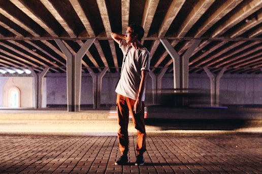 A person stands under a large structure with a complex network of beams and columns. The person is wearing a white shirt and orange pants, and appears to be relaxed, with one hand behind their head. A car is captured in motion, creating a blurred effect as it passes by the figure.